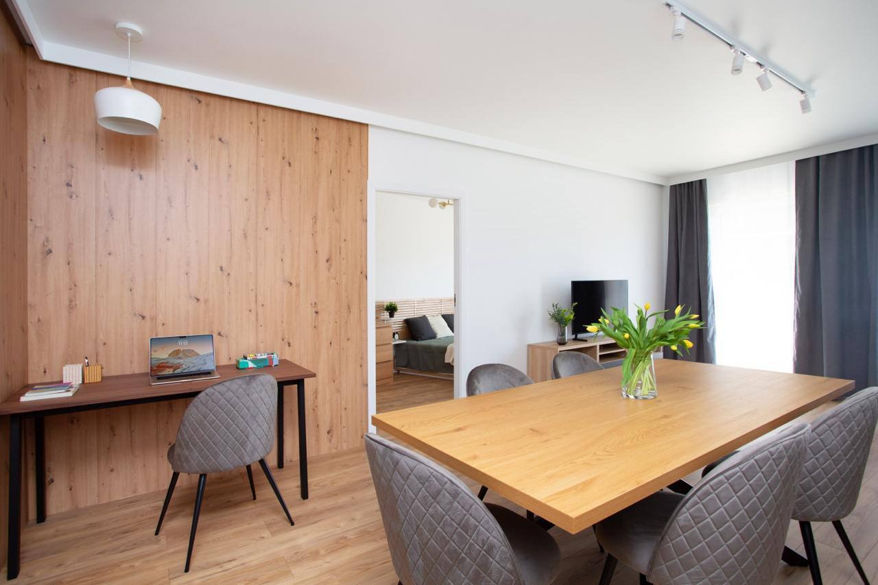 Jasne wnętrze: stół z kwiatami, biurko z laptopem pod drewnianą ścianą. Widok na sypialnię. Szare krzesła, parkiet. Nowoczesne oświetlenie.