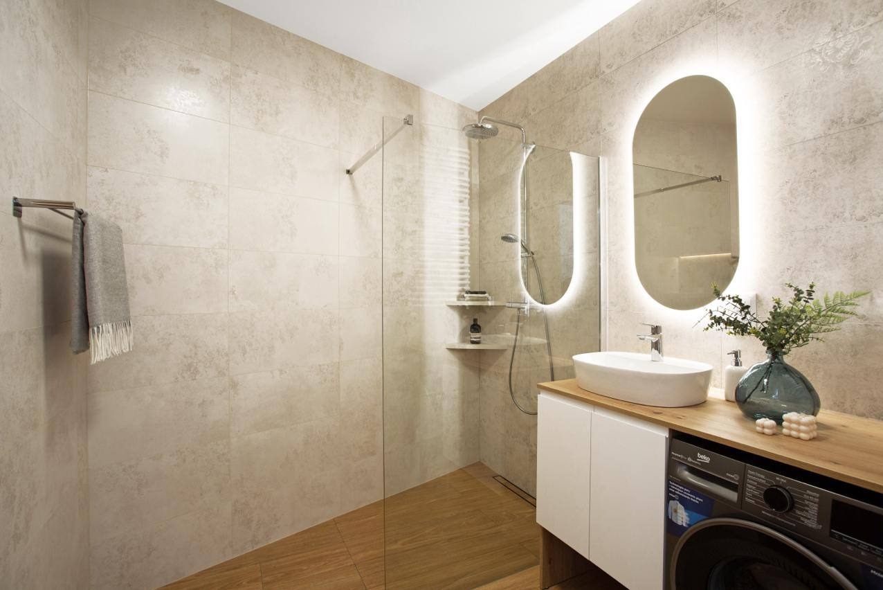 Nowoczesna łazienka w Krakowie z kabiną typu walk-in, podświetlanym lustrem i płytkami imitującymi kamień. Uwagę zwraca pralka w zabudowie.