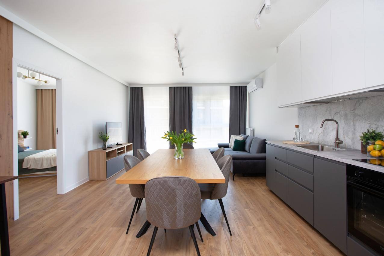 Jasne wnętrze mieszkania z drewnianą podłogą: stół z krzesłami, aneks kuchenny z marmurowym blatem, szara sofa i widok na sypialnię w tle.