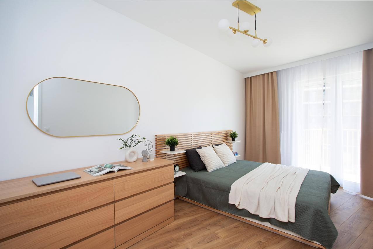 Jasna sypialnia w stylu skandynawskim: drewniane meble, łóżko z zagłówkiem z lameli, lustro w złotej ramie, lampa sufitowa i drewniana podłoga.