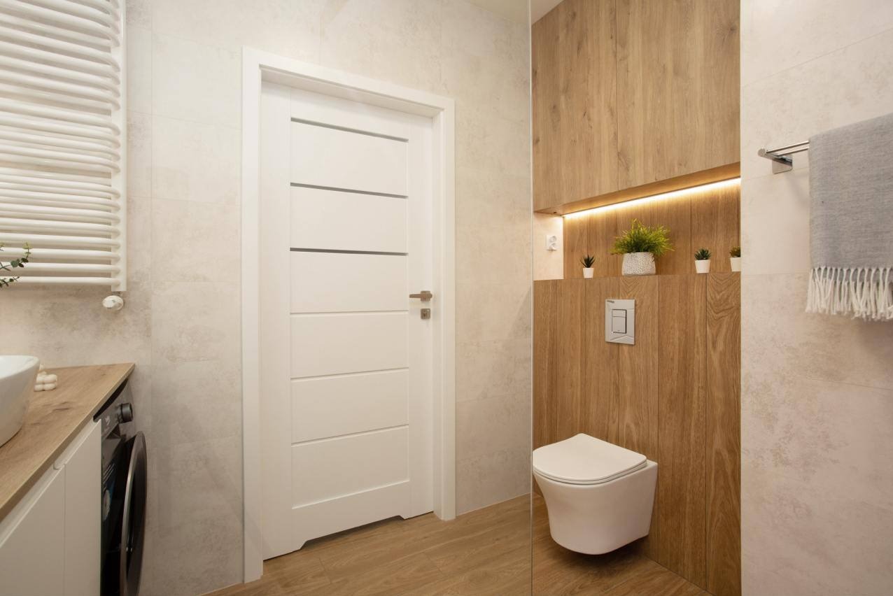 Nowoczesna łazienka z białą armaturą, drewnianą zabudową WC z oświetleniem LED i białą, minimalistyczną umywalką z pralką. Jasne płytki i białe drzwi z poziomymi frezami.