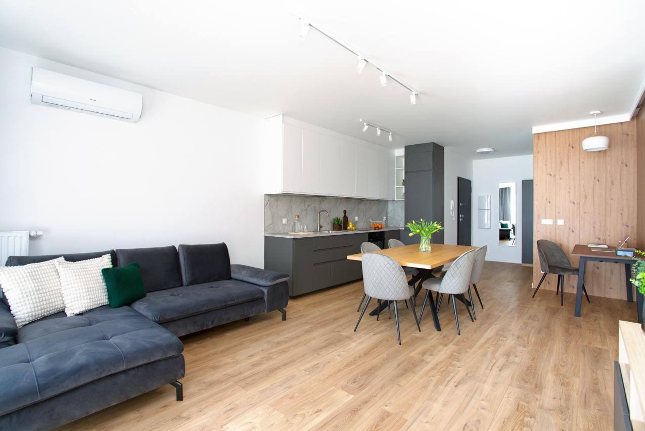 Jasne wnętrze: szary narożnik, drewniana podłoga, stół z kwiatami, kuchnia z szarymi szafkami i marmurowym blatem, białe ściany, designerskie lampy sufitowe.