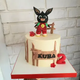 Tort urodzinowy z figurką królika Binga w czerwonej kraciastej sukience, drewnianym płotkiem i napisem 'Kuba' oraz czerwoną cyfrą '2', umieszczony na białym tle z ceglaną ścianą.