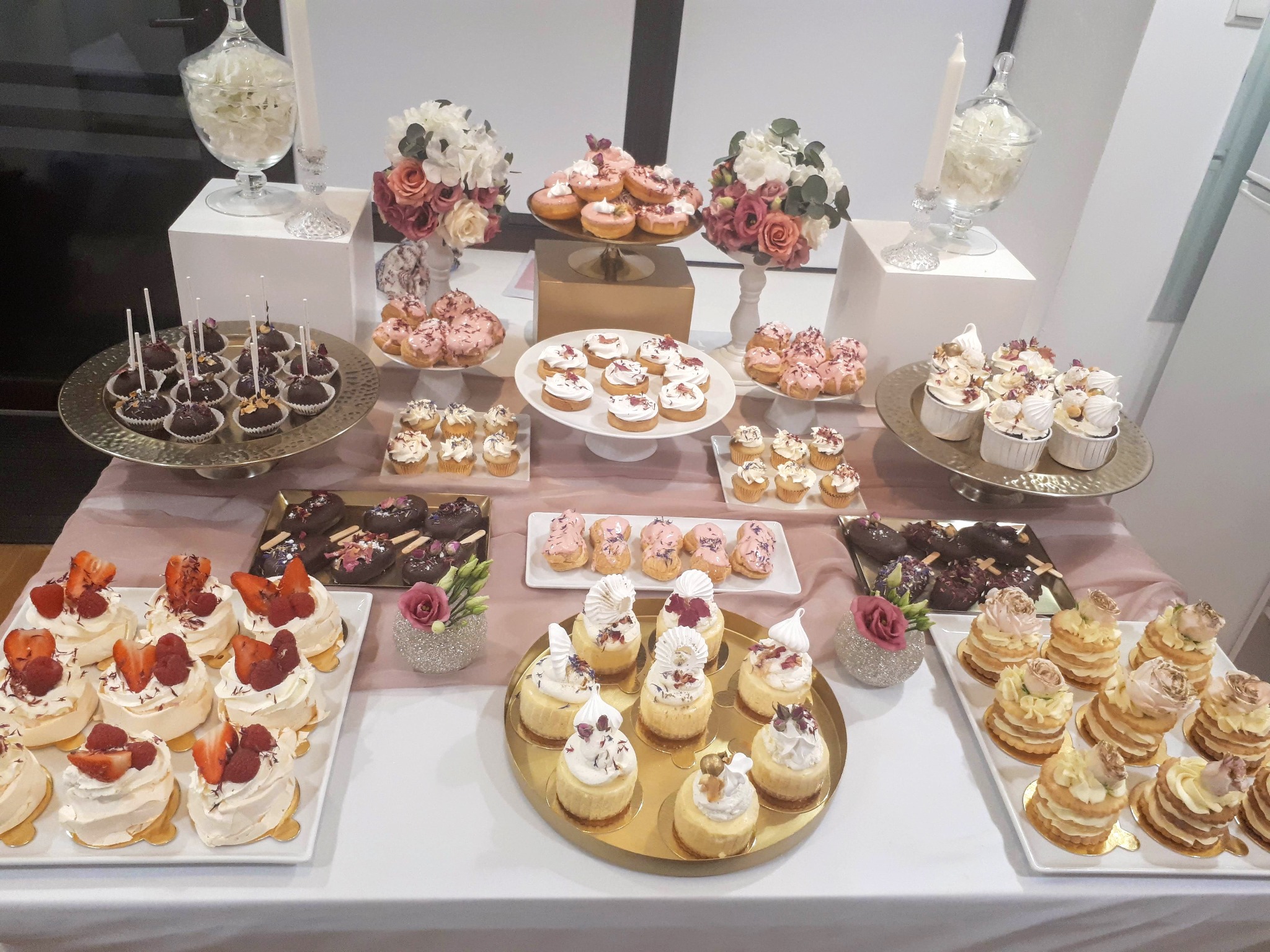 Dekoracyjny stół z różnorodnymi ciastami, babeczkami i deserami na paterach, ozdobiony kwiatami i świecami, przygotowany na uroczystość.