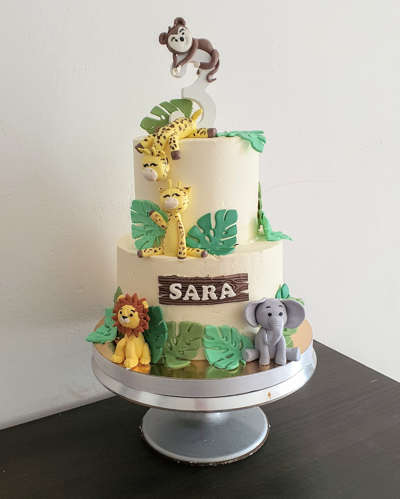 Dwupoziomowy tort urodzinowy z motywem safari, ozdobiony figurkami lwa, słonia, żyrafy, małpy i zielonymi liśćmi, z napisem 'SARA' na drewnianej tabliczce.
