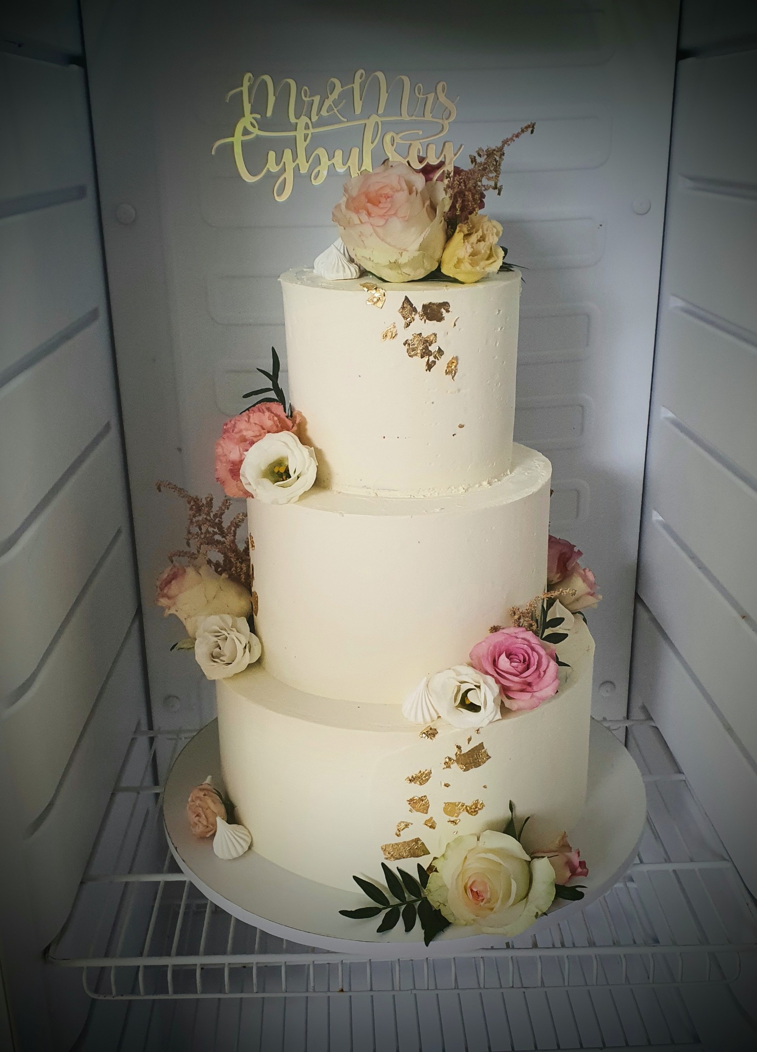 Trzypiętrowy tort weselny z białym kremem, zdobiony świeżymi różami w odcieniach różu i bieli, złotymi płatkami i topperem 'Mr & Mrs Cybula', umieszczony w chłodnym wnętrzu lodówki.