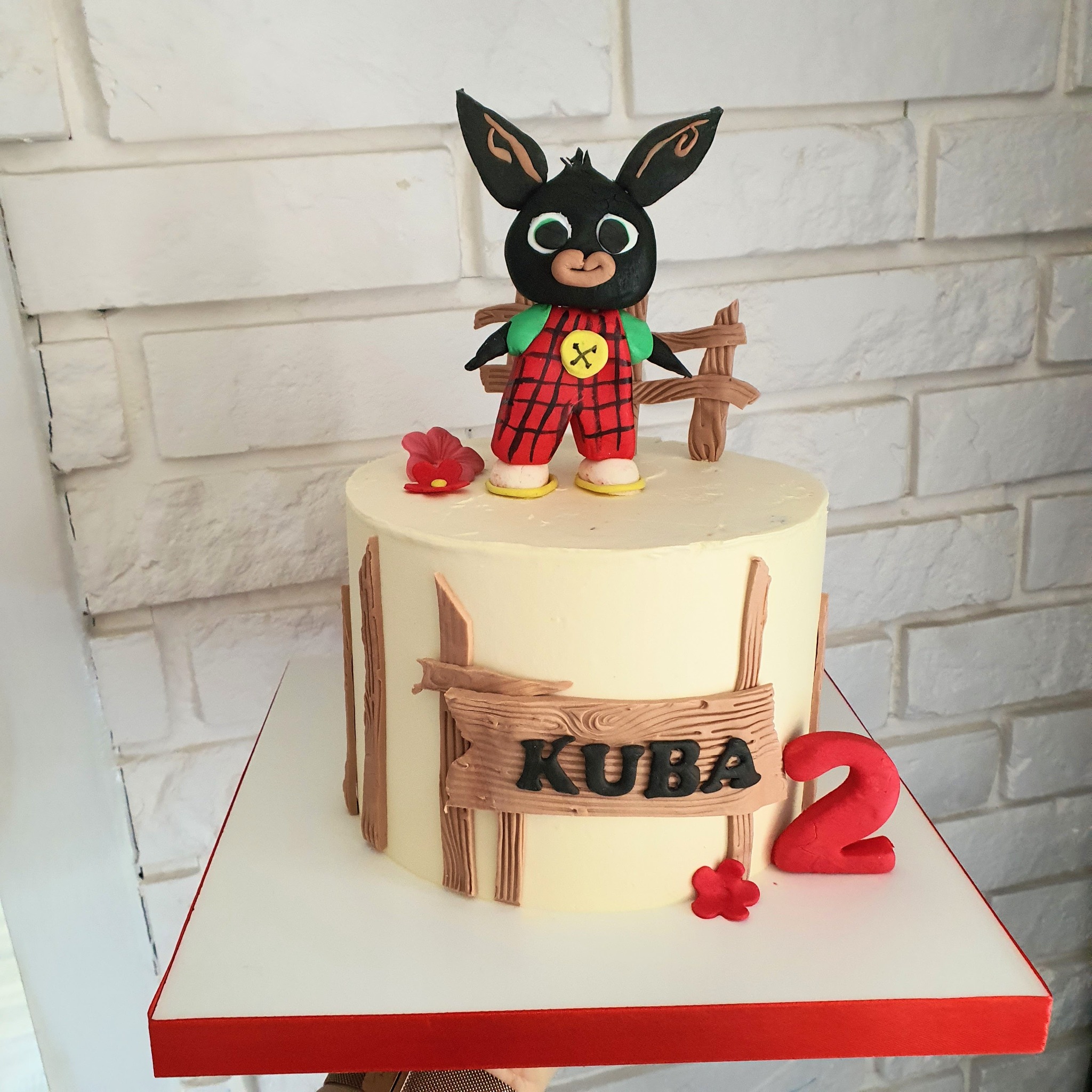 Tort urodzinowy z figurką królika Binga w czerwonej kraciastej sukience, drewnianym płotkiem i napisem 'Kuba' oraz czerwoną cyfrą '2', umieszczony na białym tle z ceglaną ścianą.