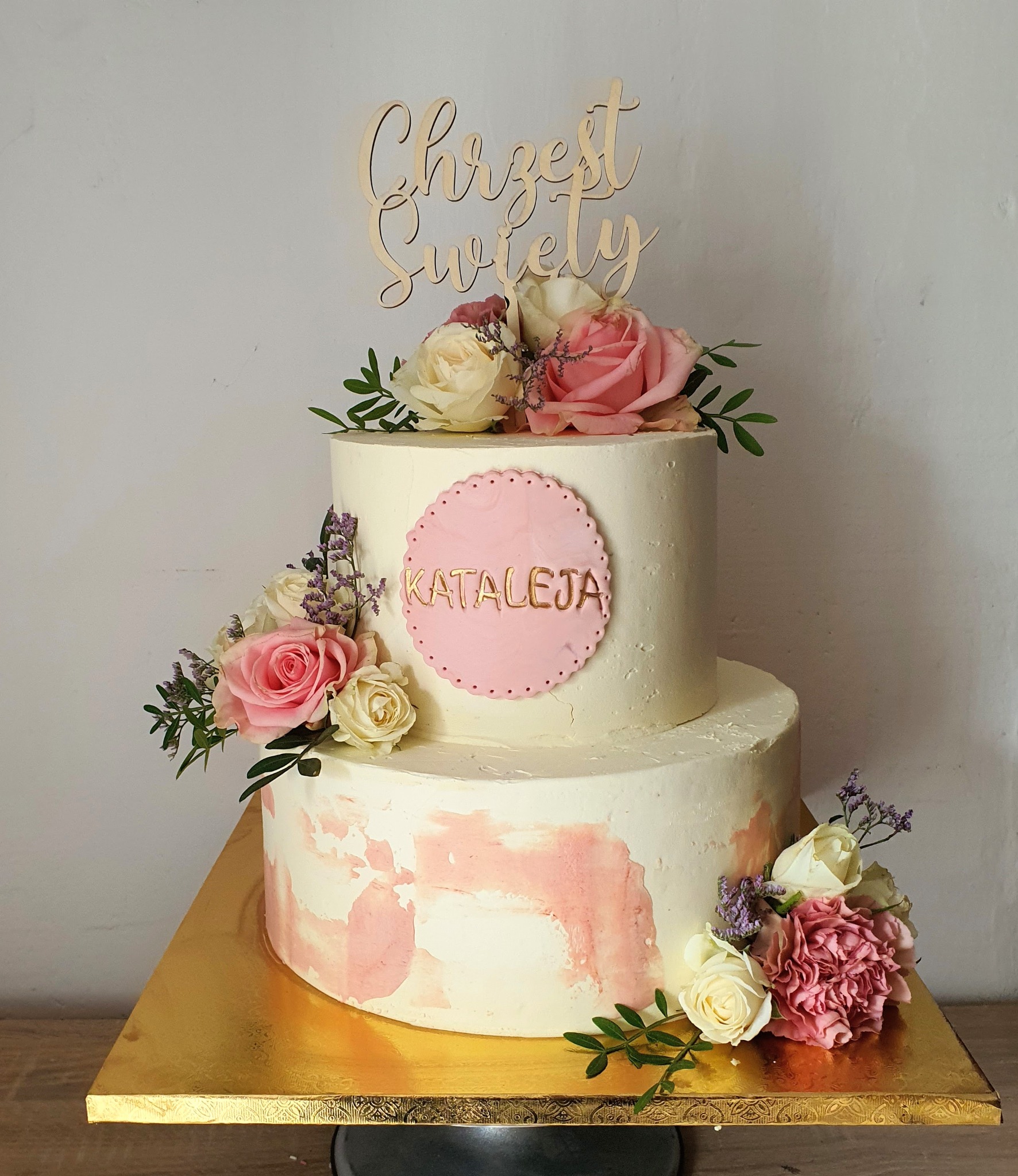 Dwupoziomowy tort z białym kremem i różowym marmurkowym wzorem, udekorowany różami, zielonymi liśćmi i topperem z napisem 'Chrzest Święty' oraz imieniem 'Katarzyna' na różowym tle.