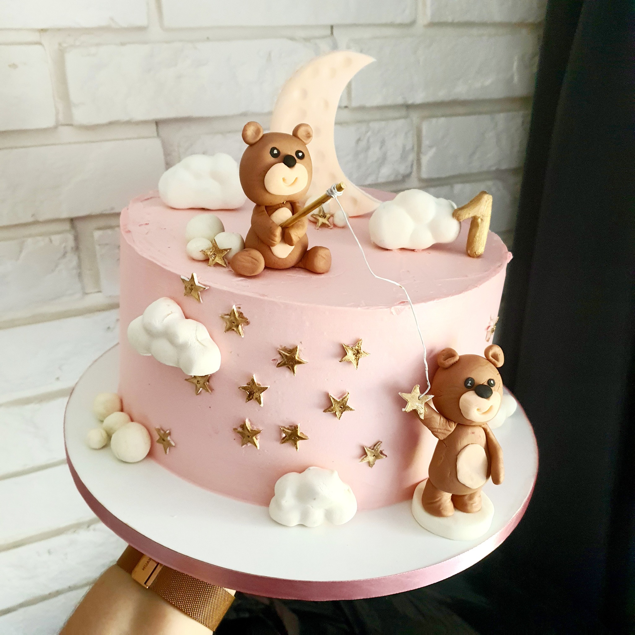 Różowy tort urodzinowy z dekoracjami: figurki misiów łowiących gwiazdy na tle księżyca i chmurek, posypany złotymi gwiazdkami, trzymany przez osobę z zegarkiem.