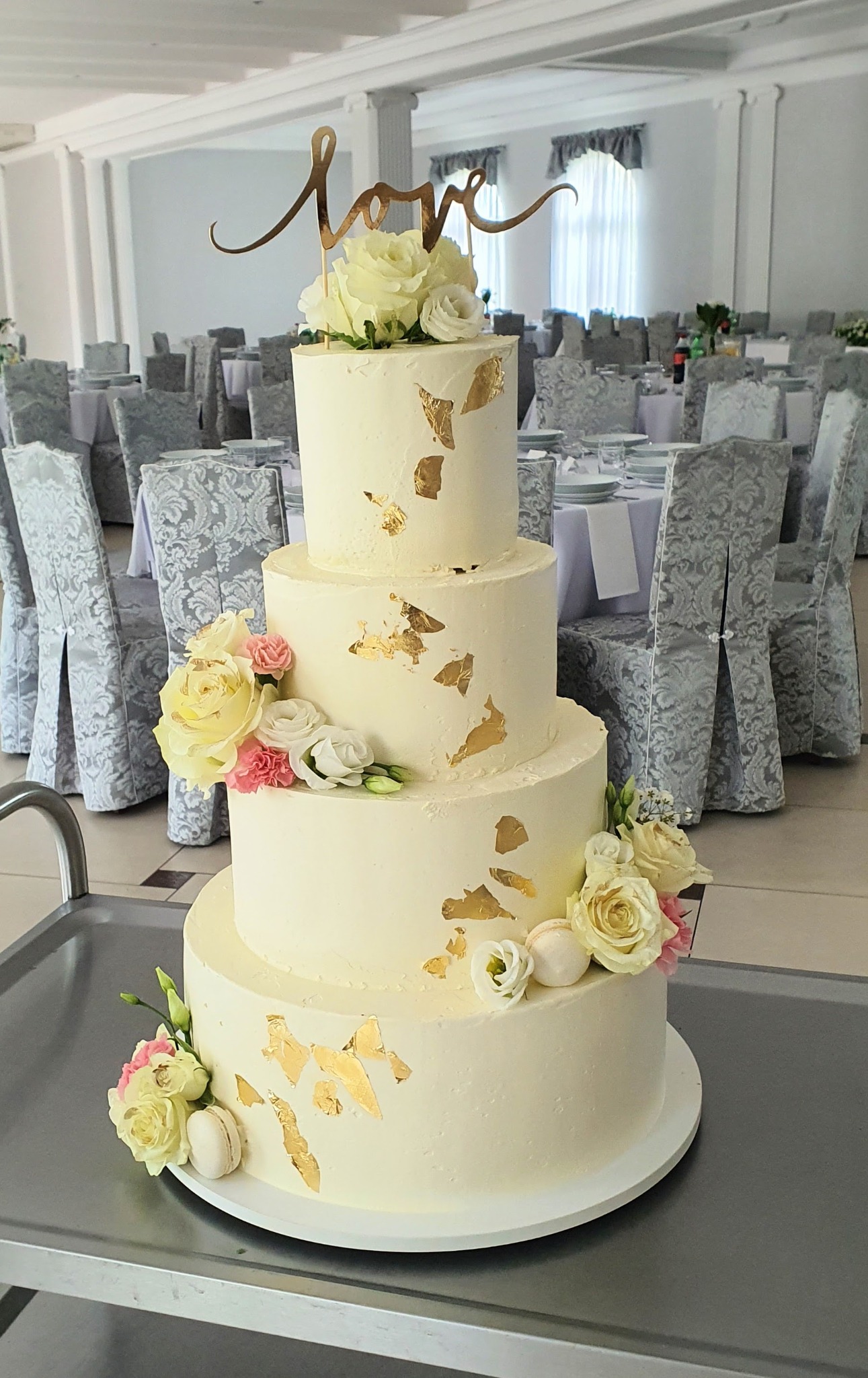Czteropiętrowy tort weselny z białym kremem, złotymi płatkami i kwiatową dekoracją na tle sali bankietowej z nakrytymi stołami i krzesłami.