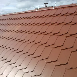 Nowe, brązowe dach&oacute;wki karpi&oacute;wki ułożone na dachu. Widoczne zabezpieczenia przeciwśniegowe i fragment nieba z chmurami. Dach widziany pod kątem.