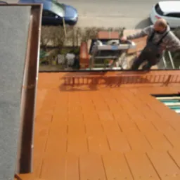 Montaż brązowych płyt na dachu domu. Widoczna drabina i pracownik w kombinezonie. Ujęcie z góry, z widokiem na częściowo pokryty dach i elementy konstrukcyjne.