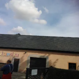 Czarny, nowy dach z dachówki na budynku z napisem 'Rebolandia'. Widok na całą połać dachu, błękitne niebo z chmurami w tle. Częściowo widoczna osoba i ogrodzenie.