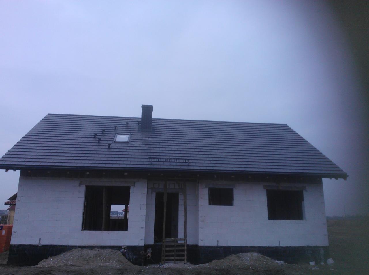 Dom w trakcie budowy z ciemną dachówką ceramiczną i oknem dachowym. Widoczny komin i niewykończone ściany z pustaków. Na dachu zamontowane elementy wentylacyjne.