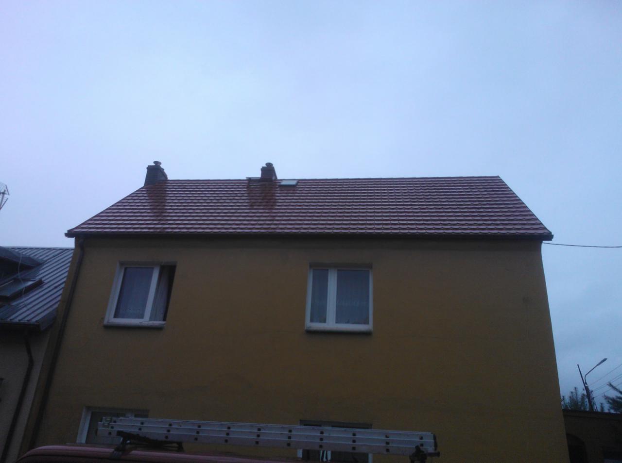 Dwukondygnacyjny budynek z nowym, brązowym pokryciem dachowym z dachówki ceramicznej. Widoczna drabina oparta o budynek. Pochmurne niebo w tle.