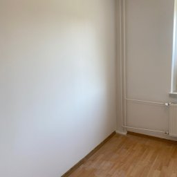 OM - Pomalowane na biało ściany w pustym pokoju z parkietem i grzejnikiem, widoczny kontakt elektryczny i lampa na suficie.