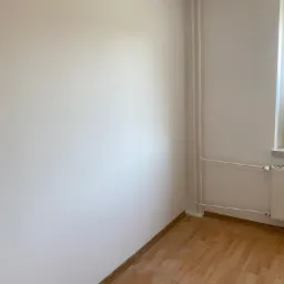 Pomalowane na biało ściany w pustym pokoju z parkietem i grzejnikiem, widoczny kontakt elektryczny i lampa na suficie.