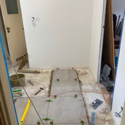 OM - Układanie szarych płytek podłogowych w trakcie remontu, widoczne narzędzia glazurnicze, poziomica, system poziomowania płytek i wiadro z zaprawą.