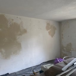 OM - Pokój w trakcie prac przygotowawczych do wykończenia: ściany z miejscowymi nierównościami po nałożeniu zaprawy, profile metalowe i narzędzia na podłodze zabezpieczonej folią, okno z widokiem...