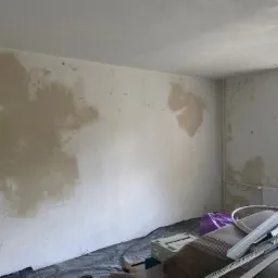 Pok&oacute;j w trakcie prac przygotowawczych do wykończenia: ściany z miejscowymi nier&oacute;wnościami po nałożeniu zaprawy, profile metalowe i narzędzia na podłodze zabezpieczonej folią, okno z widokiem...