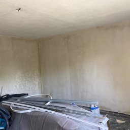 OM - Wnętrze pokoju w trakcie prac wykończeniowych, ściany i sufit pokryte świeżo nałożoną, nierówną warstwą, widoczne narzędzia pracy i odkurzacz budowlany.
