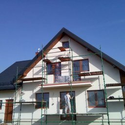 OM - Dom jednorodzinny w trakcie prac wykończeniowych elewacji, widoczne rusztowania, pracownik z wiadrem farby, okna zabezpieczone folią, błękitne niebo.