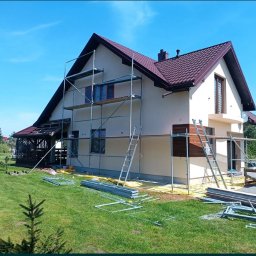 OM - Dom jednorodzinny w trakcie malowania elewacji, widoczne rusztowania, zabezpieczona folią strefa przy budynku, częściowo pomalowana fasada w kolorze kremowym, brązowe elementy dekoracyjne, zielony...