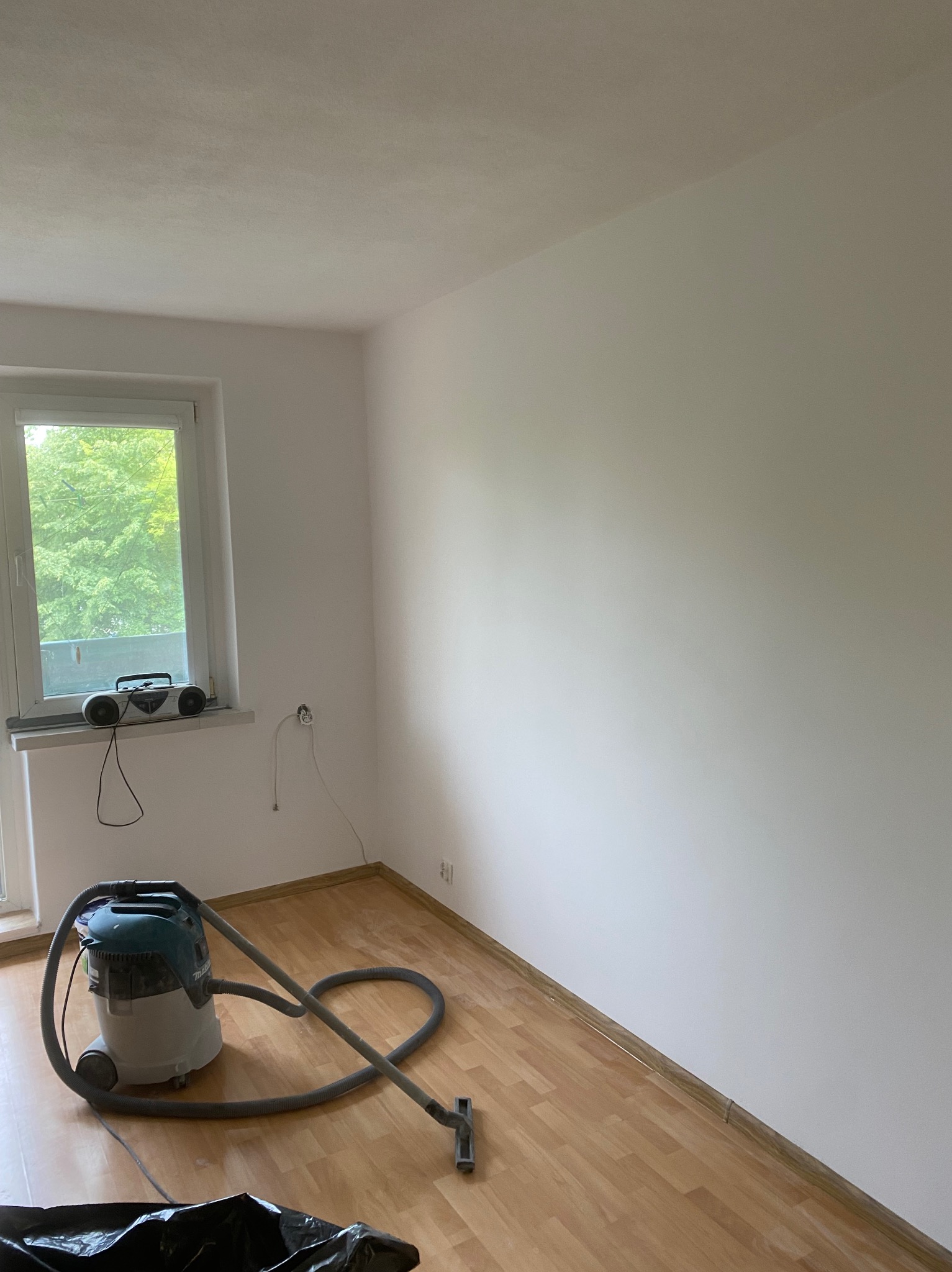 Pomalowane na biało wnętrze pokoju z parkietem, widoczny odkurzacz przemysłowy Makita, radio na parapecie i gniazdko elektryczne.