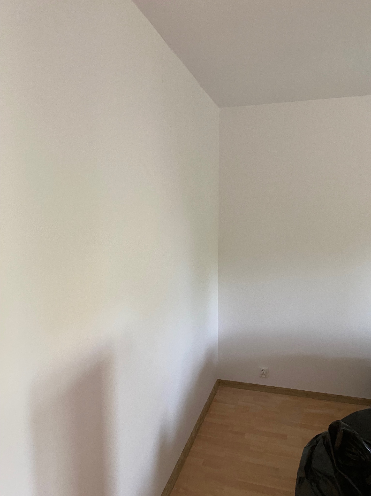 Świeżo pomalowany narożnik pokoju z gładkimi, białymi ścianami i drewnianą podłogą; widoczne gniazdko elektryczne i fragment czarnej folii ochronnej na podłodze.