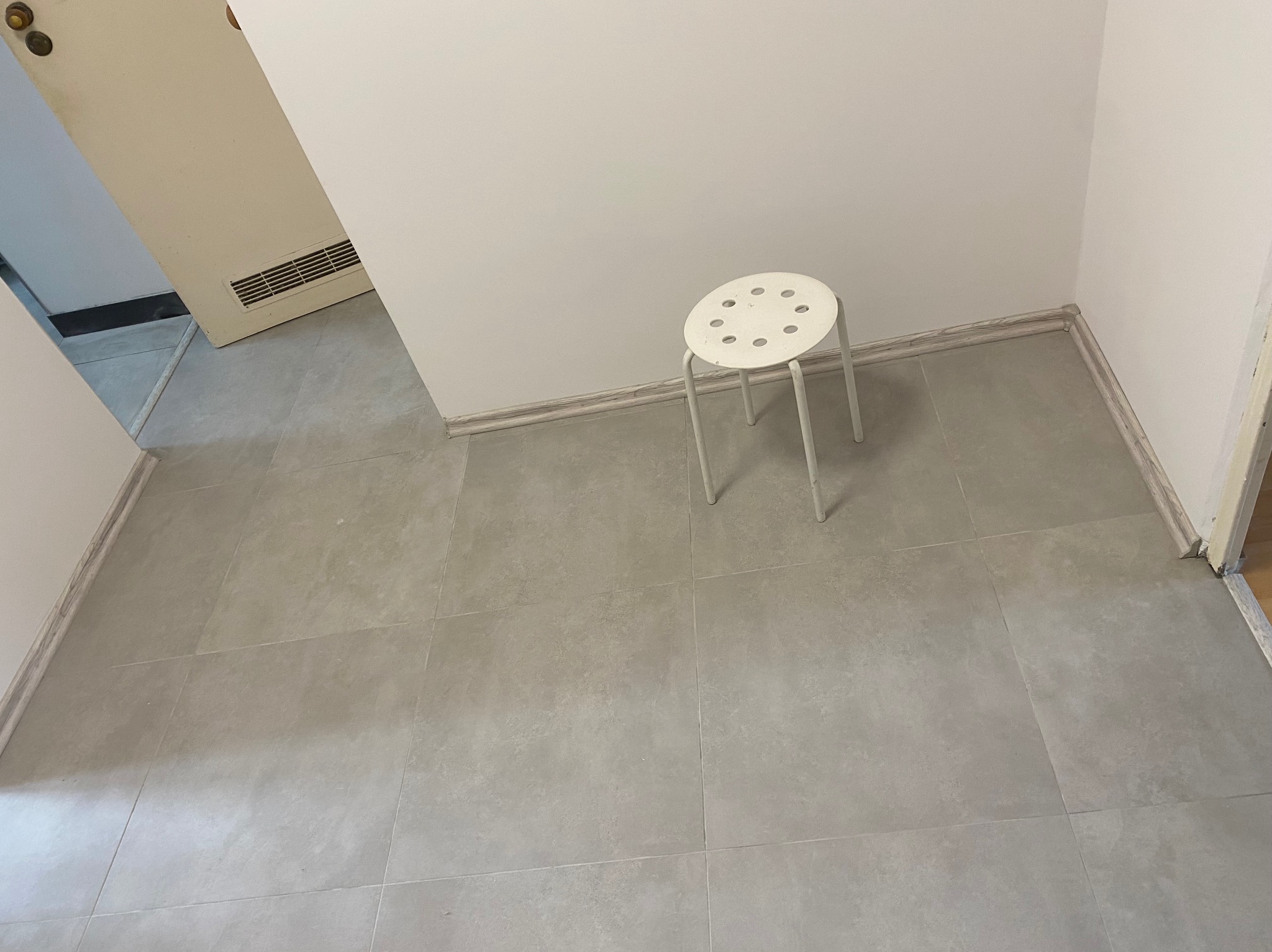 Świeżo położona, szara, matowa podłoga z płytek w pustym pokoju, widoczny biały taboret i fragment otwartych drzwi.