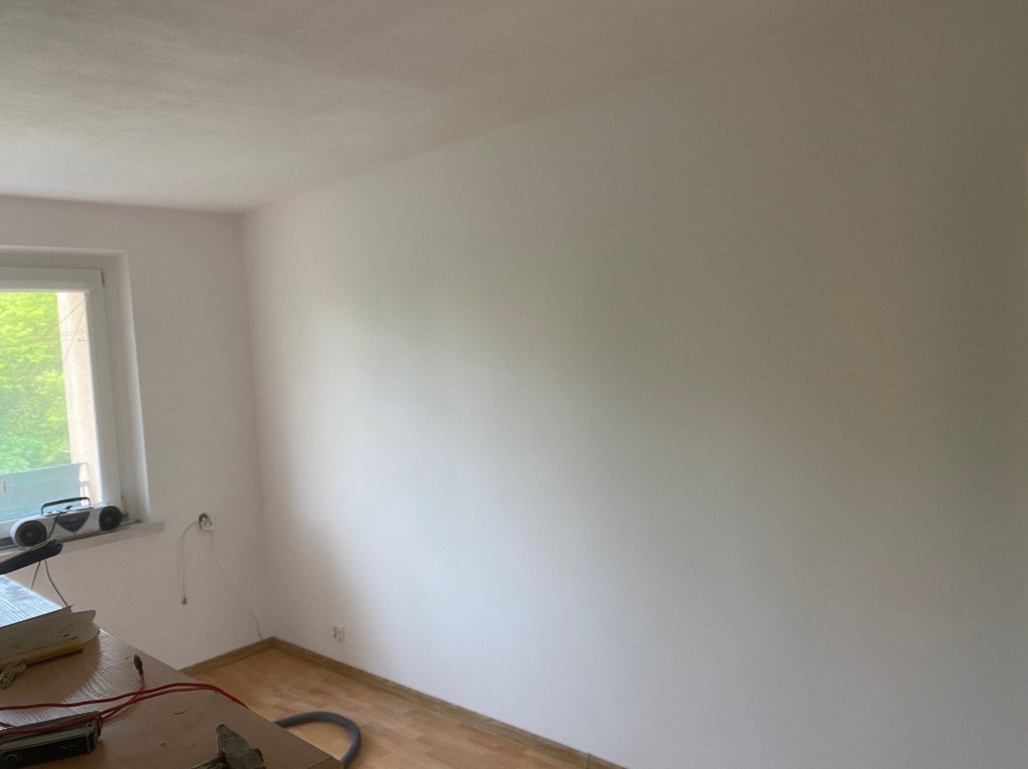 Pomalowane na biało ściany w pokoju z drewnianą podłogą, widoczne gniazdko elektryczne wystające ze ściany, oraz narzędzia na stole w rogu pomieszczenia.