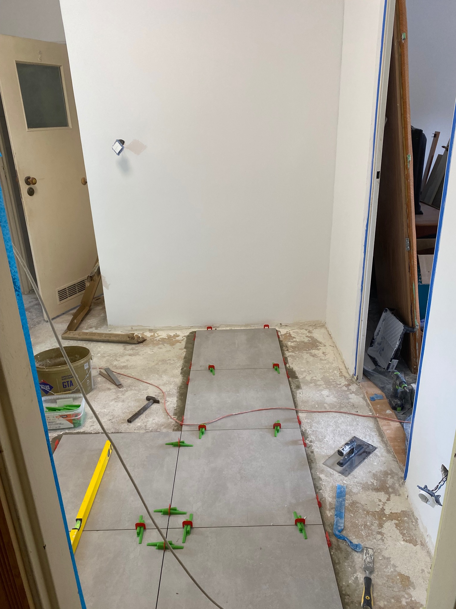 Układanie szarych płytek podłogowych w trakcie remontu, widoczne narzędzia glazurnicze, poziomica, system poziomowania płytek i wiadro z zaprawą.