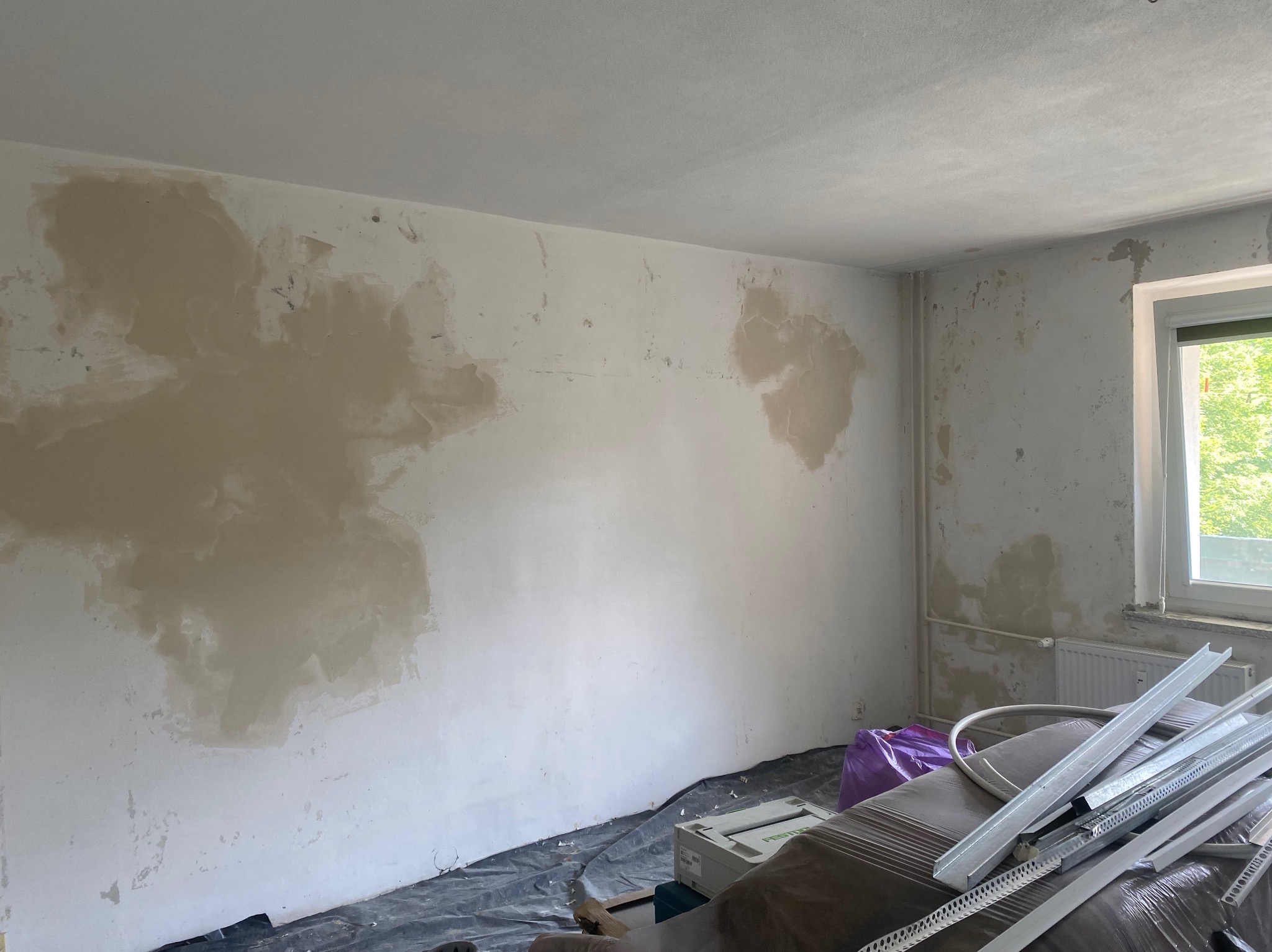 Pokój w trakcie prac przygotowawczych do wykończenia: ściany z miejscowymi nierównościami po nałożeniu zaprawy, profile metalowe i narzędzia na podłodze zabezpieczonej folią, okno z widokiem...