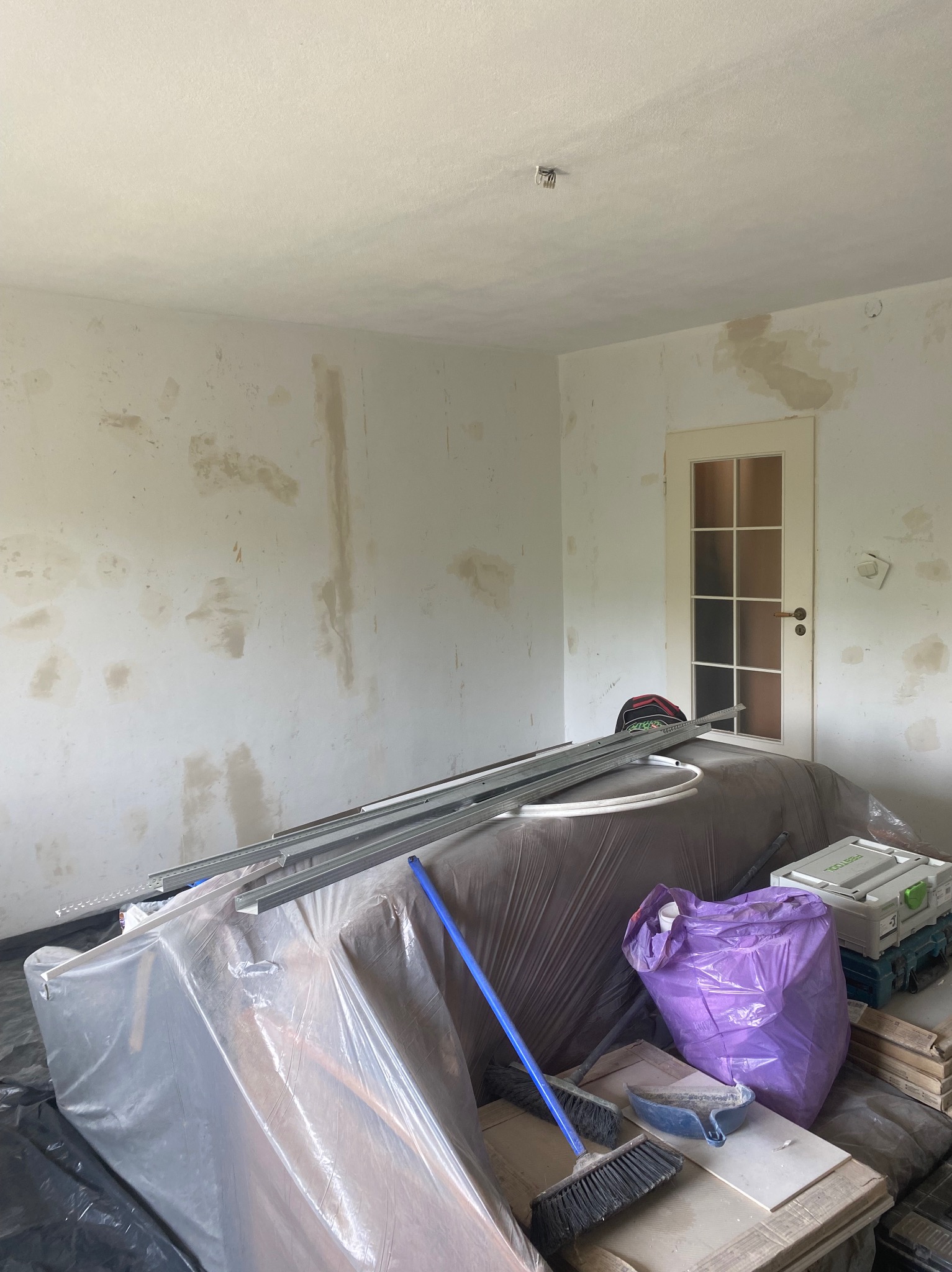 Pokój w trakcie przygotowań do prac wykończeniowych: ściany ze śladami po łataniu, zabezpieczone meble folią, widoczne narzędzia i materiały, drzwi z szybami w tle.