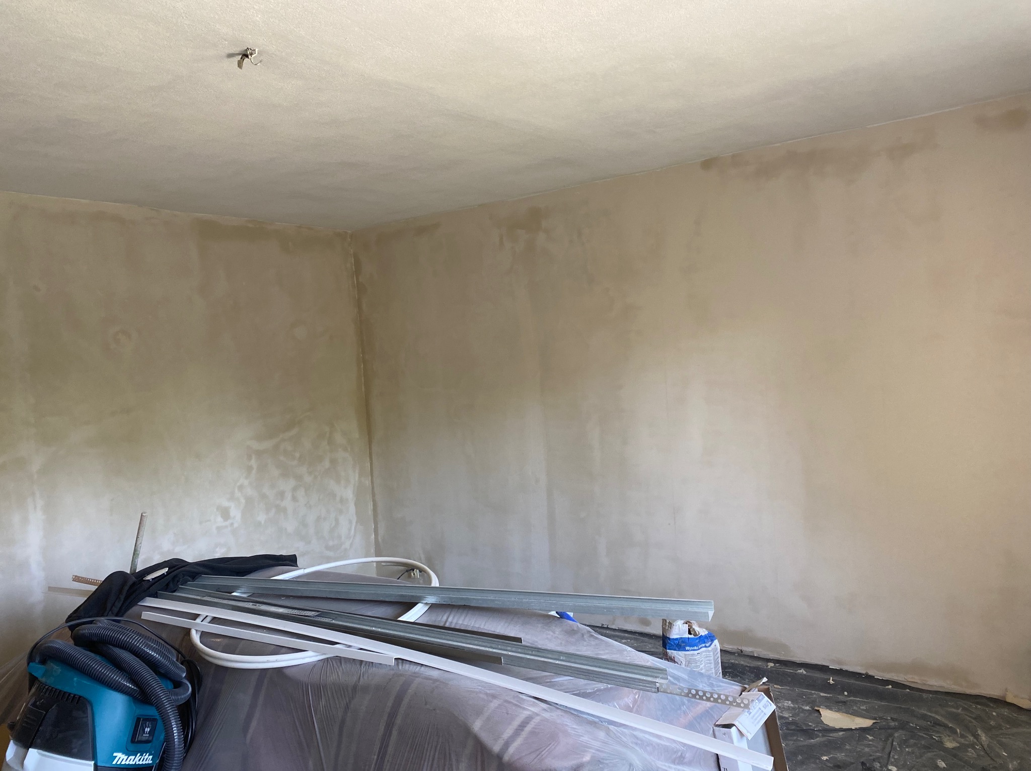 Wnętrze pokoju w trakcie prac wykończeniowych, ściany i sufit pokryte świeżo nałożoną, nierówną warstwą, widoczne narzędzia pracy i odkurzacz budowlany.