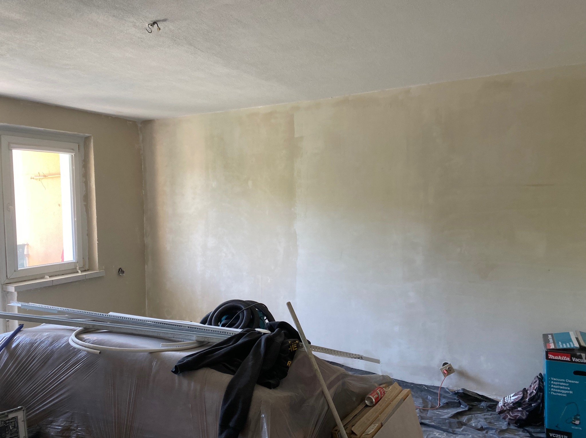 Pokój w trakcie prac wykończeniowych: ściany pokryte świeżą warstwą, widoczne narzędzia i materiały budowlane zabezpieczone folią, okno z naturalnym światłem.