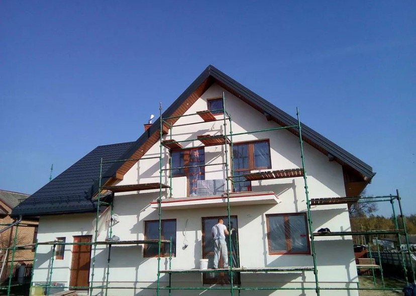 Dom jednorodzinny w trakcie prac wykończeniowych elewacji, widoczne rusztowania, pracownik z wiadrem farby, okna zabezpieczone folią, błękitne niebo.