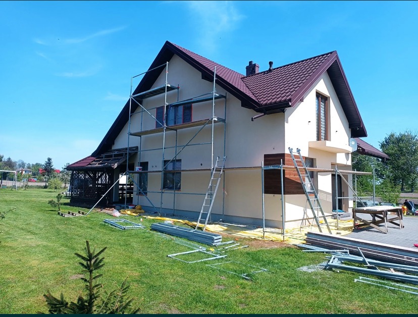 Dom jednorodzinny w trakcie malowania elewacji, widoczne rusztowania, zabezpieczona folią strefa przy budynku, częściowo pomalowana fasada w kolorze kremowym, brązowe elementy dekoracyjne, zielony...