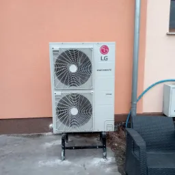 Zewnętrzna jednostka dwuwentylatorowej pompy ciepła LG Therma V, zamontowana na metalowej podstawie przy budynku w Zielonej Górze, obok rury spustowej i plecionej sofy.