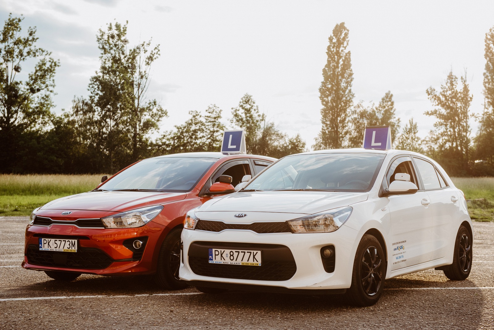 Dwa samochody Kia Rio: czerwony i biały, z niebieskimi znakami 'L' na dachu, zaparkowane na parkingu z drzewami w tle.