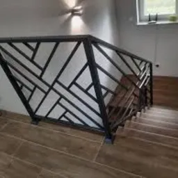 Nowoczesna, metalowa balustrada schodowa z geometrycznym wzorem, czarne profile na jasnym tle ściany, widok z góry na drewniane schody.