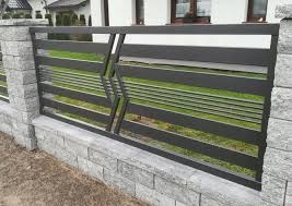 Nowoczesne przęsło ogrodzeniowe z poziomych profili stalowych w kolorze antracytowym, z ukośnym wzorem w centralnej części, zamontowane na murku z szarej kostki łupanej. W tle trawnik i fragment domu.