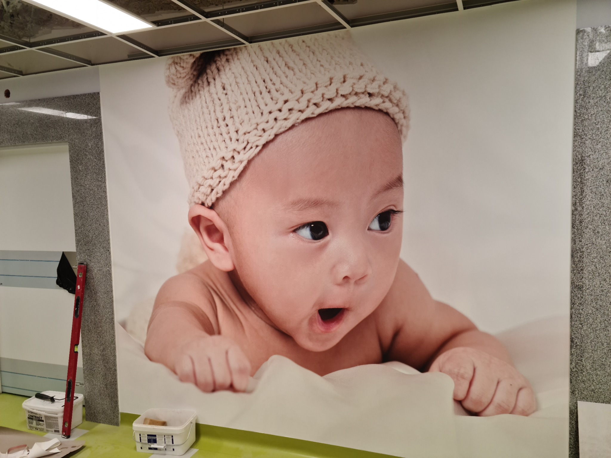 Świeżo położona fototapeta z dużym zdjęciem niemowlęcia w wełnianej czapce, widoczne narzędzia i poziomica przy ścianie.
