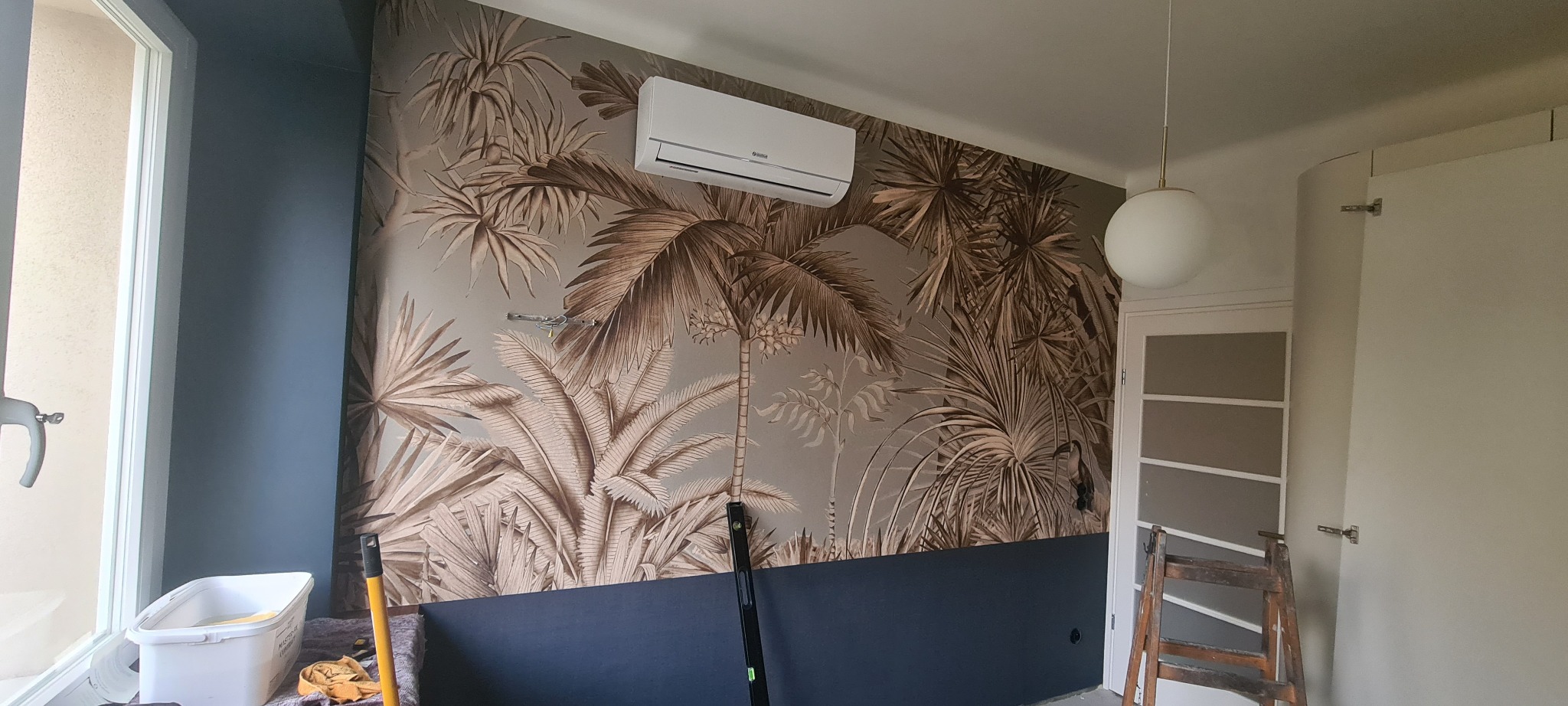 Pokój po remoncie z tapetą w motywy tropikalne, widoczny klimatyzator, wiadro z klejem, drabina i poziomica.