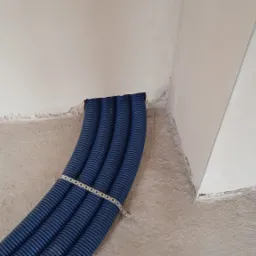 Niebieskie rury karbowane, spięte taśmą perforowaną, wystają ze ściany w surowym stanie, widoczne niedociągnięcia w tynku przy otworze.