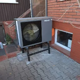 Szara pompa ciepła marki Heiko zamontowana na metalowym stelażu przy ścianie budynku z cegły, widoczne okno piwniczne i fragment elewacji z brązową boazerią.