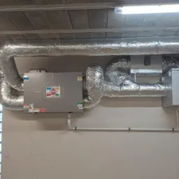 Szara jednostka centralna rekuperacji z aluminiowymi, izolowanymi przewodami wentylacyjnymi, zamontowana pod betonowym stropem w pomieszczeniu z oknem. Widoczne białe rury i przewody elektryczne.