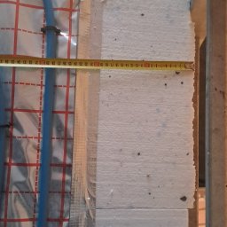 Marek Gomułka GOKIMA Engineering - Ułożone niebieskie rury ogrzewania podłogowego na srebrnej macie izolacyjnej z czerwoną siatką, obok białych płyt styropianu, z żółtą miarką mierzącą szerokość izolacji.