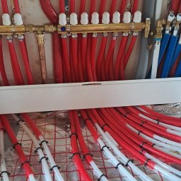 Marek Gomułka GOKIMA Engineering - Mosiężny rozdzielacz 'PERFEKT' z białymi pokrętłami, podłączony do czerwonych rur w osłonach i białych rur systemu ogrzewania podłogowego, ułożonych na izolacji.
