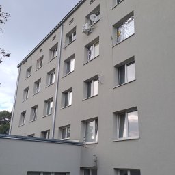 Marek Gomułka GOKIMA Engineering - Jasnoszara elewacja wielopiętrowego budynku z zamontowaną jednostką klimatyzacji zewnętrznej i anteną satelitarną. Widoczne otwarte okna, niektóre z folią termoizolacyjną.